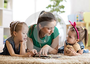 Young mother reading a book to her kids daughters. Children and mom lying on rug in sunny living room.