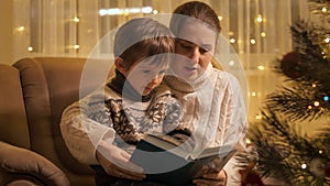 Young mother reading book to her son sitting in armchair next to glowing Christmas tree. Families and children