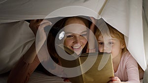 Young mother reading goodnight story fairytale to child daughter under duvet blanket in night room