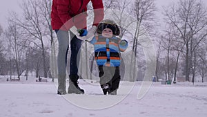 Young mother in red down jacket helps her little baby boy to make his first steps and they walking together to the