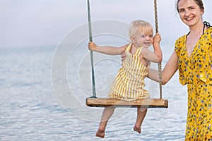 Young mother rolls the child on swing at the sea. Mom and child at the sea look at the camera and smile