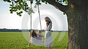 A young mother shakes her daughters on a wooden rope swing.