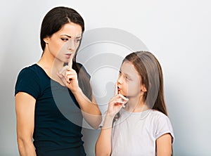 Young mother showing the finger the silence sign holding the finger near the mouth to her cute serious daugther on blue background