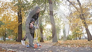 Young mother showing her toddler son how to ride a scooter in a autumn park. Active family leisure. Training, sports