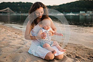 Young mother sitting on the beach with one year old baby son. Boy hugging, smiling, laughing, summer day. Happy childhood carefree