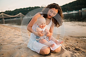 Young mother sitting on the beach with one year old baby son. Boy hugging, smiling, laughing, summer day. Happy childhood carefree