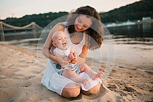 Young mother sitting on the beach with one year old baby son. Boy hugging, smiling, laughing, summer day. Happy childhood carefree