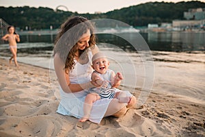 Young mother sitting on the beach with one year old baby son. Boy hugging, smiling, laughing, summer day. Happy childhood carefree