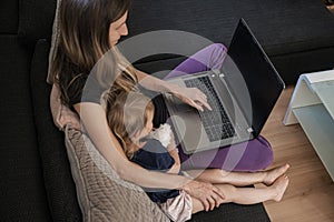 Young mother sitting on the couch working on laptop computer with her daughter next to her