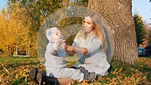 Young mother sitting on the grass with her little baby, smiling. Waving hand to the cameraman.