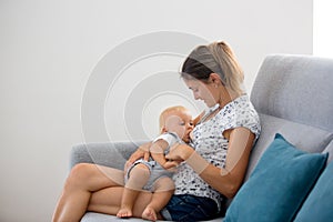 Young mother, sitting at home in sunny living room, cuddling with her toddler baby boy, breastfeeding him