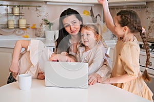 Young mother sitting with laptop on kitchen and three daughters interfere her work