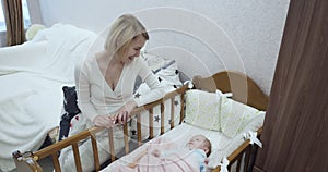 Young mother sitting near the crib looks on her newborn baby.