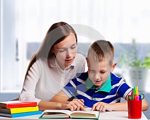 Young mother sitting at a table at home helping her small son with his homework from school as he writes notes in a notebook