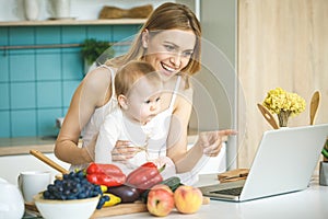 Young mother smiling, cooking and playing with her baby daughter in a modern kitchen. Using laptop. Healthy food concept.