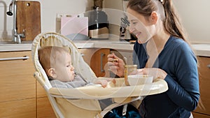 Young mother smiling at her baby son sitting in highchair at kitchen. Concept of parenting, healthy nutrition and baby care