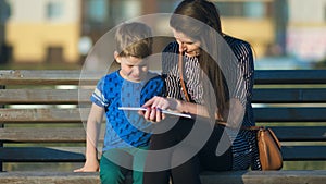 Young mother and son sitting on a bench in a park and reading a book