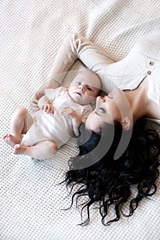 Young mother spends time with her newborn son, playing together, lying on a white bed in the bedroom