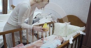Young mother stands near the crib shows yellow toy to newborn baby.