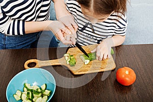 Young mother teaches her little daughter to cut cucumber for healthy salad