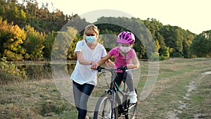 A young mother teaches his daughter to ride a Bicycle in medical masks and a Bicycle helmet near the river. Protection FROM covid-