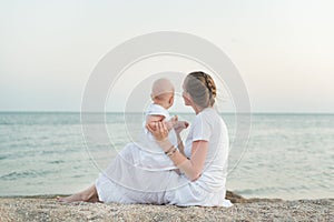 Young mother and toddler sitting on the sandy shore of sea background. Motherhood, harmony and love