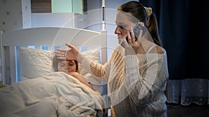 Young mother touching forehead of her sick son lying in bed an talking to doctor by phone. Concept of children illness