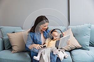 Young mother trying to entertain her newborn baby by playing cartoons and funny videos on digital tablet while sitting on sofa at