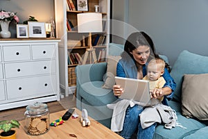 Young mother trying to entertain her newborn baby by playing cartoons and funny videos on digital tablet while sitting on sofa at