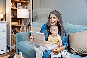 Young mother trying to entertain her newborn baby by playing cartoons and funny videos on digital tablet while sitting on sofa at