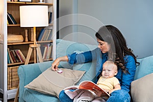 Young mother trying to entertain her newborn baby by reading a fairytale books while sitting on sofa at home. Woman read a book