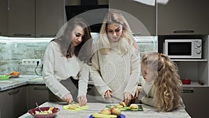 Young mother and two her daughters cooks fruits salad at kitchen. Mother and her daughters cuts an oranges for fruit