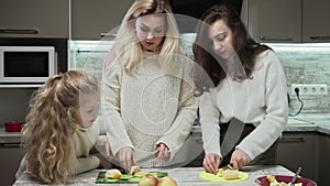 Young mother and two her daughters cooks fruits salad at kitchen. Mother and her daughters cuts an oranges for fruit