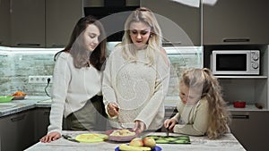 Young mother and two her daughters cooks fruits salad at kitchen. Mother mixes a fruit salat with bananas and oranges