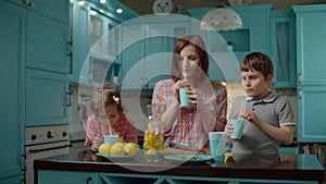Young mother with two kids drinking homemade lemonade standing on blue kitchen at home. Mom with son and daughter