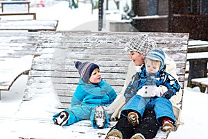Young mother with two little toddler boys sitting on bench in winter park. Woman playing and hugging with cute baby sons