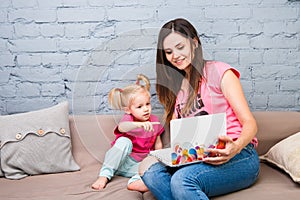 Young mother and two year old girl with blond hair sitting on the couch and using laptop. They are dressed in bright pink clothes.