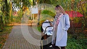 Young mother using telephone in hand and walking with baby in the stroller in park autumn. Child in a modern