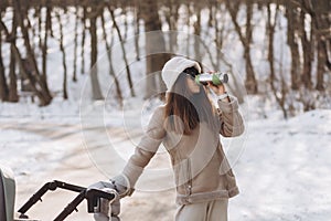 Young mother walking with baby in stroller and drinking hot coffee in winter park