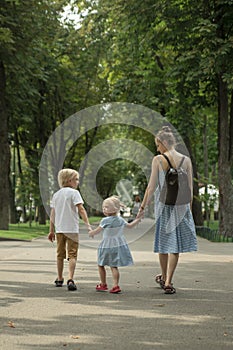 Young mother walks through summer park with her two children. Family walks along the alley in park. Back view. Vertical frame