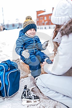 Young mother wears socks for skates, helps a little boy 3-5 years old child. In winter, on the rink in the city. Rest on
