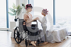 Young mother in wheelchair holds hands of her little daughter on weekend.