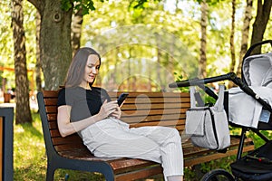 Young mother, woman with smartphone in hands sitting on Park bench next to stroller with baby, happy long-awaited motherhood