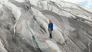 A young mountaineer in sunglasses in a cap and crampons stands on a large glacier with huge cracks in the mountains of