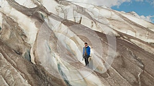 A young mountaineer in sunglasses in a cap and crampons stands on a large glacier with huge cracks in the mountains of