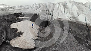 A young mountaineer in sunglasses in a cap and crampons stands on a large glacier with huge cracks in the mountains of