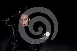 A young mulatto girl in a leather jacket and black clothes poses on an abandoned sand road. at night in the light of car