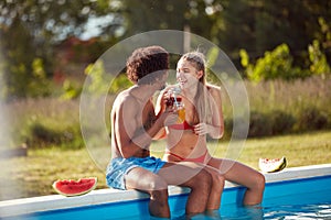 Young multiethnic couple sitting on the edge of the pool, drinking juices,  smiling, laughing