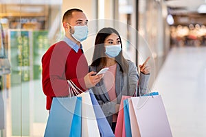 Young multiracial couple wearing face masks, walking with shopper bags at mall, checking shopping list on smartphone