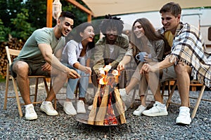 Young multiracial friends having outdoor picnic near RV, frying marshmallows on campfire, having fun time in summer